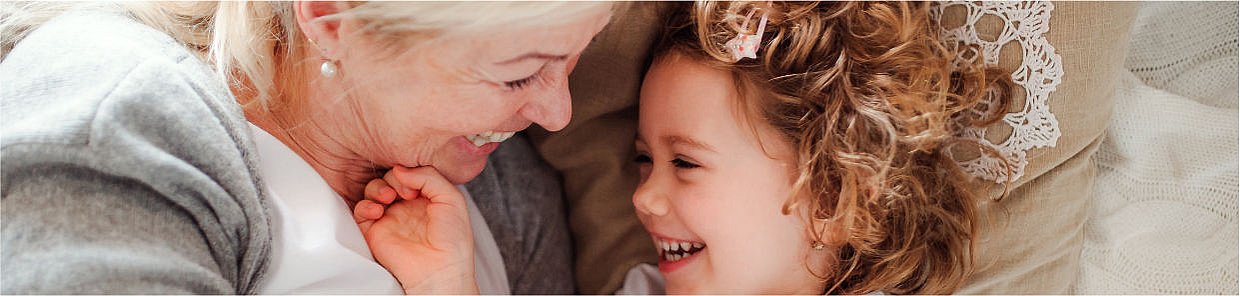 Headerbild – Fröhliche Szene auf dem Sofa: Eine ältere Frau und ein kleines Mädchen liegen eng nebeneinander und lachen sich herzlich an. Beide wirken vertraut und glücklich. Im Hintergrund eine gehäkelte Spitzen-Decke