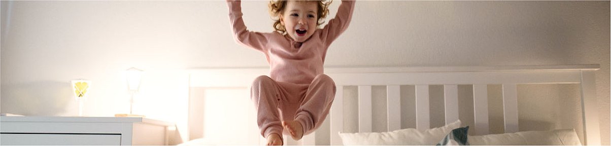 Headerbild - Kind in rosafarbener Schlafkleidung springt lachend auf dem Bett – Symbolbild für nächtlichen Husten bei Kindern und ungestörte Momente