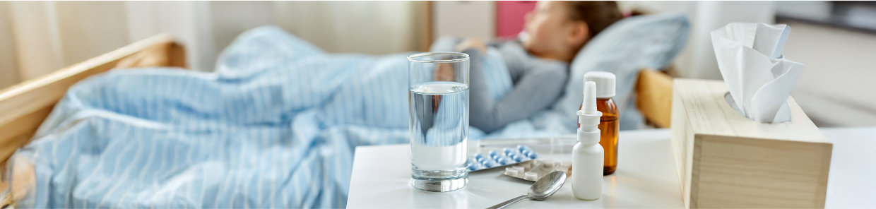 Ein Kind liegt krank im Bett mit blau-weiß gestreifter Bettwäsche. Auf dem Nachttisch stehen ein Glas Wasser, Medikamente, Nasenspray, ein Löffel und Papiertaschentücher.