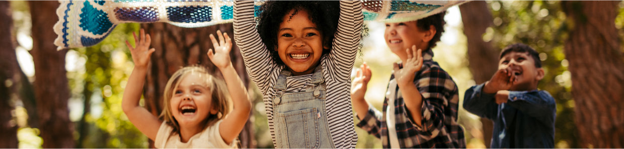 Vier Kinder spielen im Freien und halten eine bunte, gehäkelte Decke über ihren Köpfen. Im Hintergrund sind Bäume und viel Grün zu sehen, was auf einen Wald hinweist.
